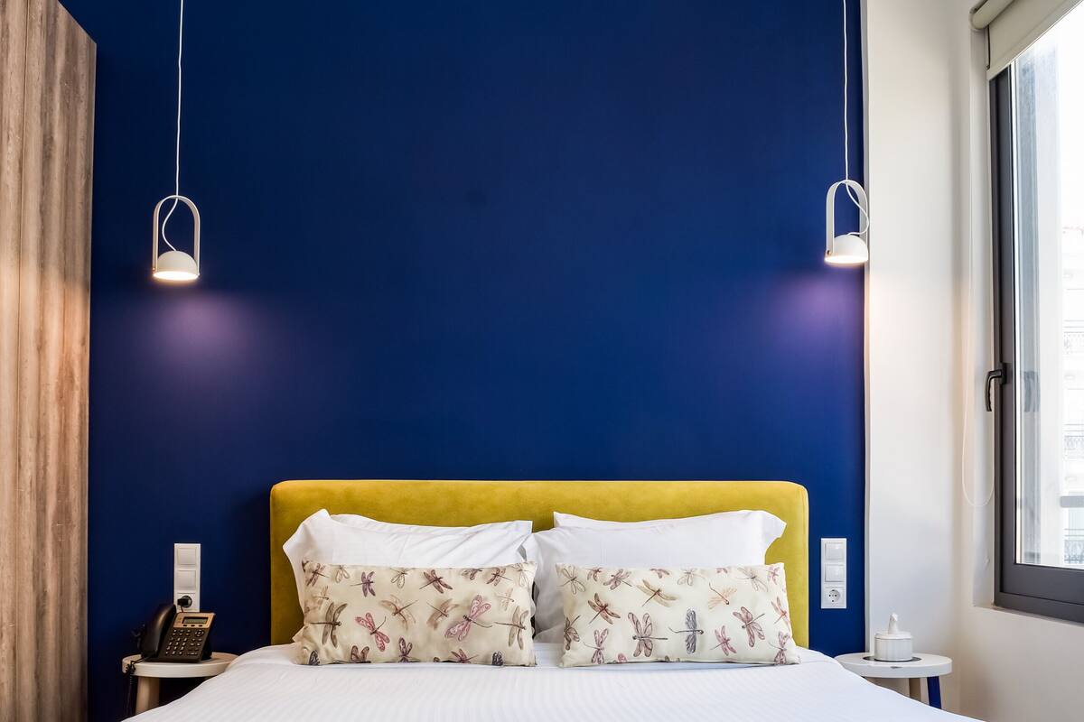 Una cama con cabecera amarilla, ropa de cama blanca y un cojín con estampado floral. Hay dos mesitas de noche, una con una lámpara y la otra con un teléfono. El piso es de madera y una pared tiene detalles azules.