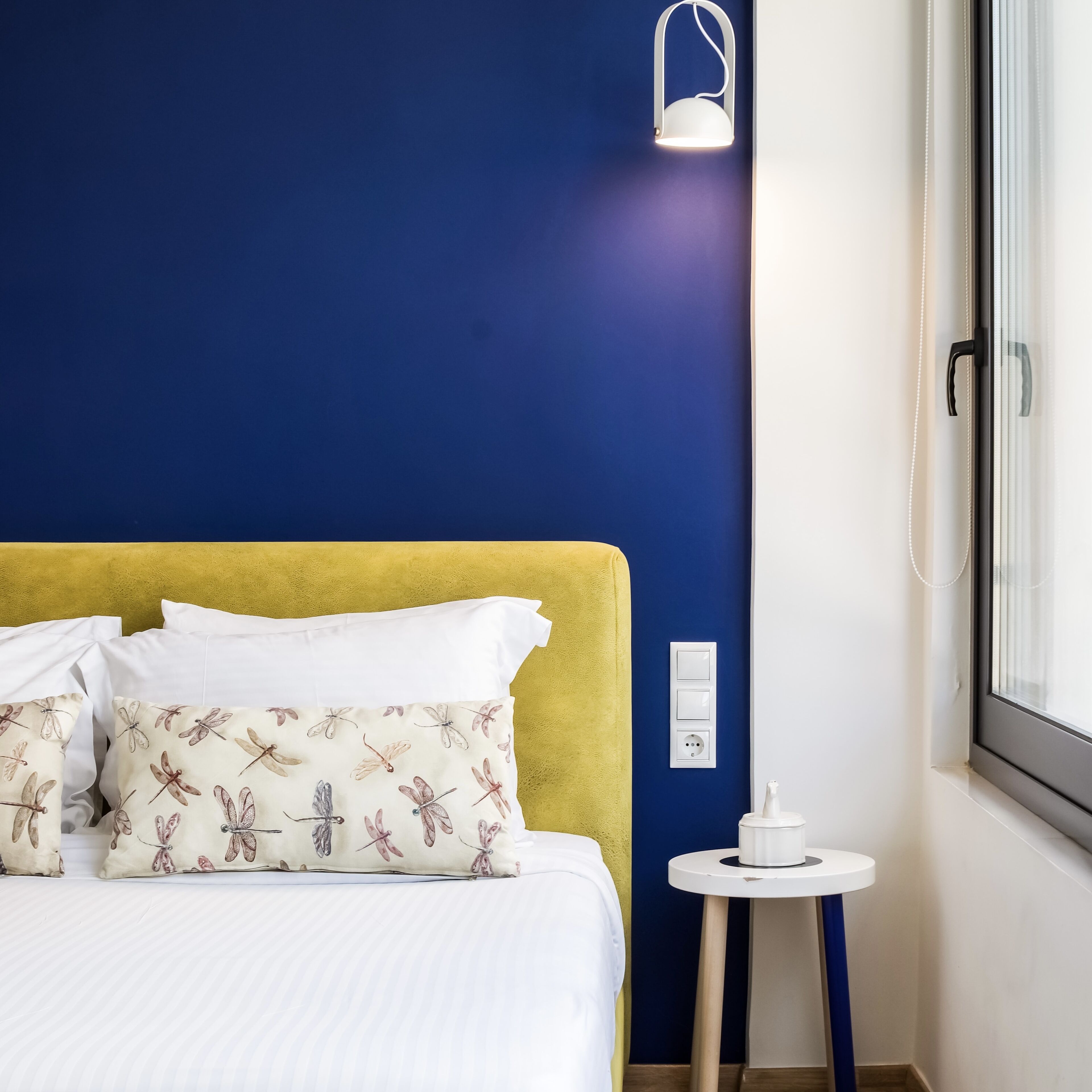 A hotel room with a bed, a bedside table, a lamp, and a window.
