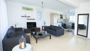 A modern living room with a sofa, TV, coffee table, and a small table with a decorative item. A kitchen is visible in the background. - Villa Sirena - Pernera (Paralimni)