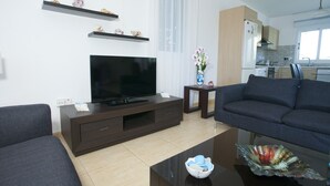 A modern living room with a flat-screen TV, dark sofas, a coffee table, and a kitchen in the background. - Villa Sirena - Pernera (Paralimni)