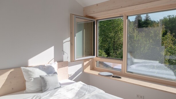 Un dormitorio con cama, almohadas, mesita de noche y ventana con vista a árboles.