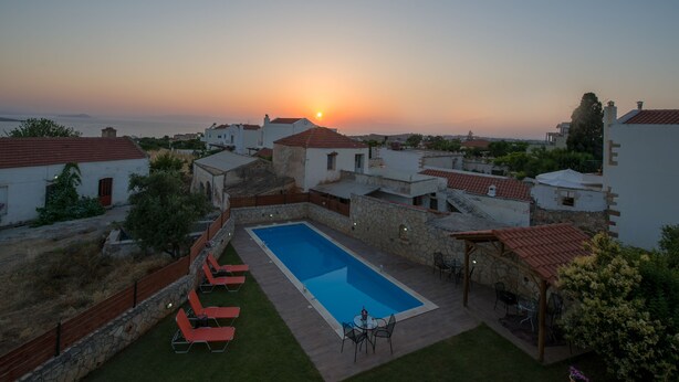 A swimming pool with lounge chairs and a table, surrounded by stone walls and red-tiled roofs, with a sunset view in the background. - Skandalis Apartment with pool view (Theano) (CHORAFAKIA CHANIA)