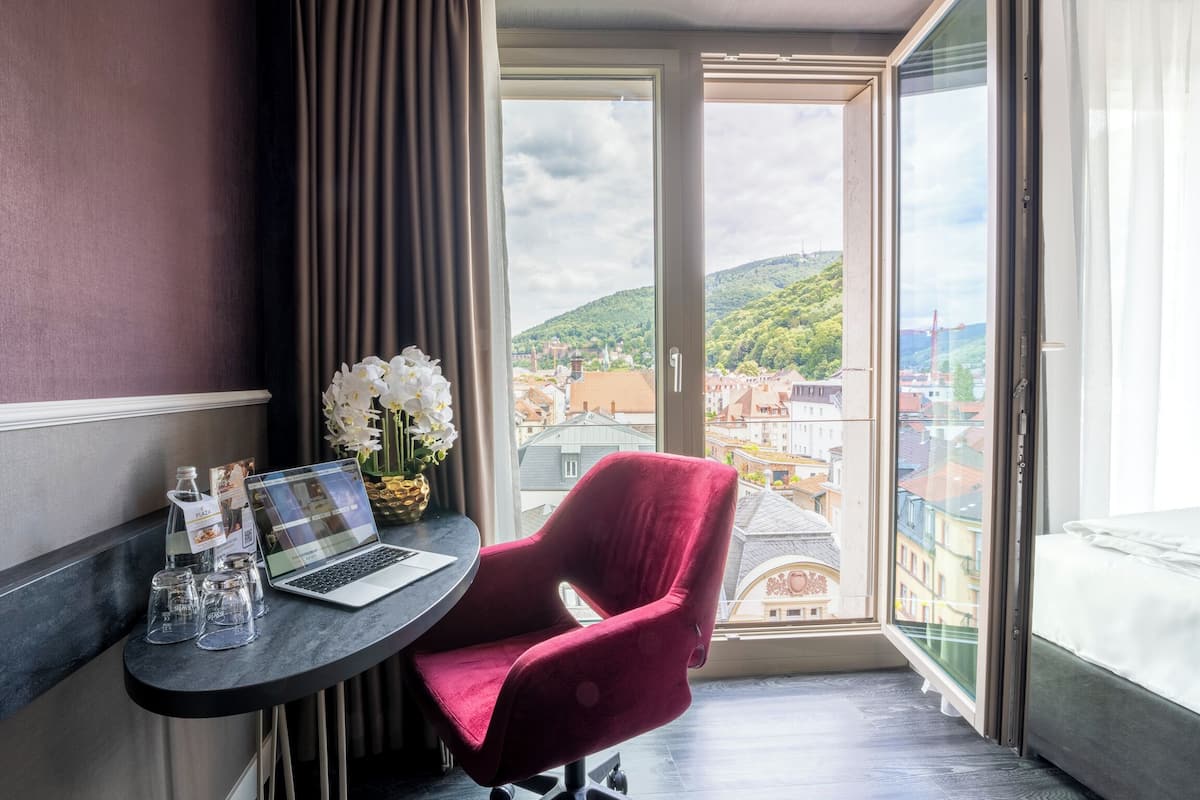 A hotel room with a bed, a desk with a laptop, a red chair, a vase with flowers, and a view of a cityscape through a large window.