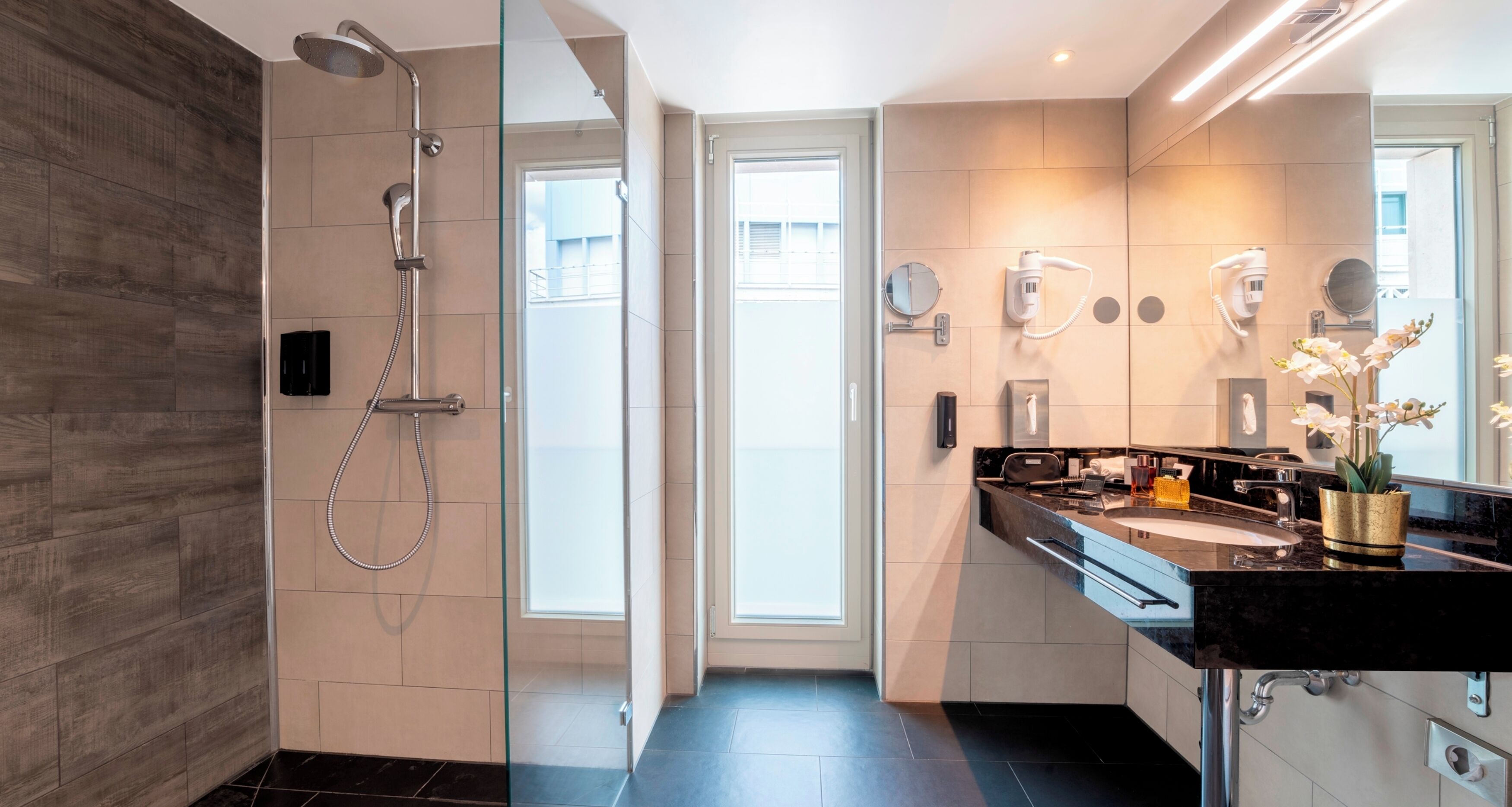 Un bagno moderno con cabina doccia in vetro, lavabo su piano nero e specchio con un altro specchio tondo sopra.