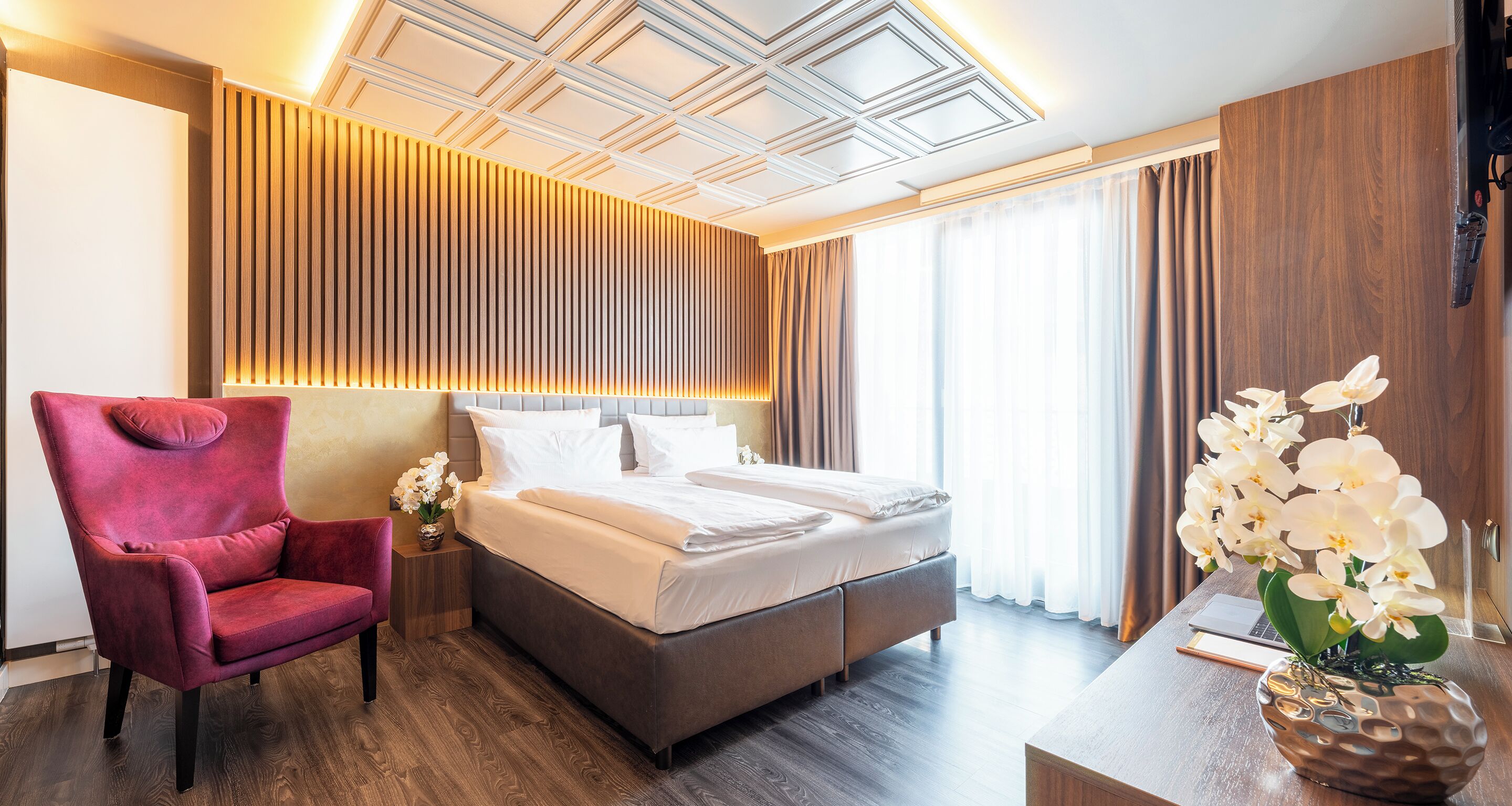 A modern hotel room with a large bed, a red armchair, a wooden headboard, and a decorative orchid vase.