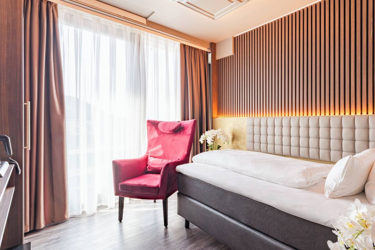 A hotel room with a bed, a red chair, a wooden panel wall, and a window with curtains.