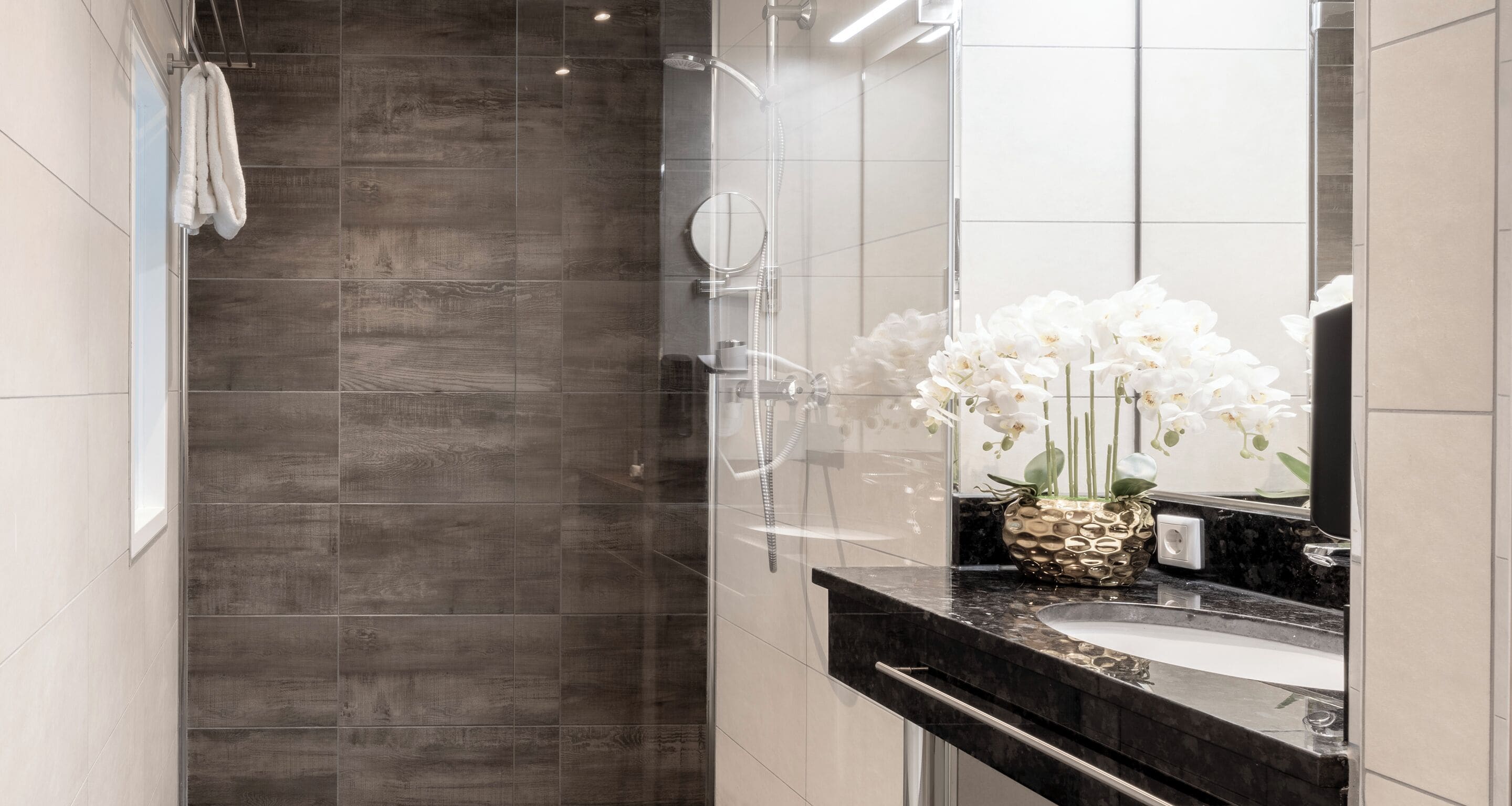 Une salle de bain moderne avec une douche aux parois en bois foncé, un grand miroir et un vase rempli d’orchidées blanches posé sur le comptoir.