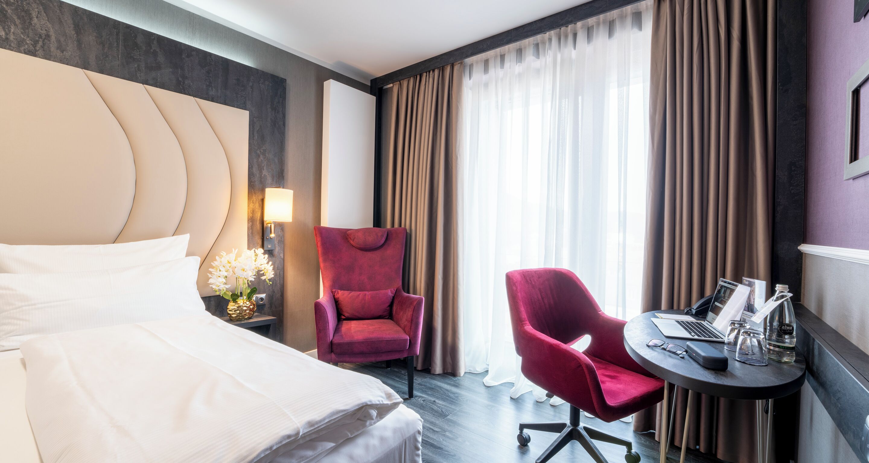 A hotel room with a bed, a red armchair, a desk with a laptop, and a window with curtains.