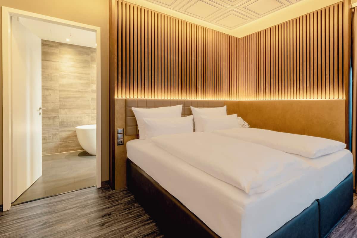 A hotel room with a large bed, a wooden headboard, a bathroom with a bathtub, and a view of the city.