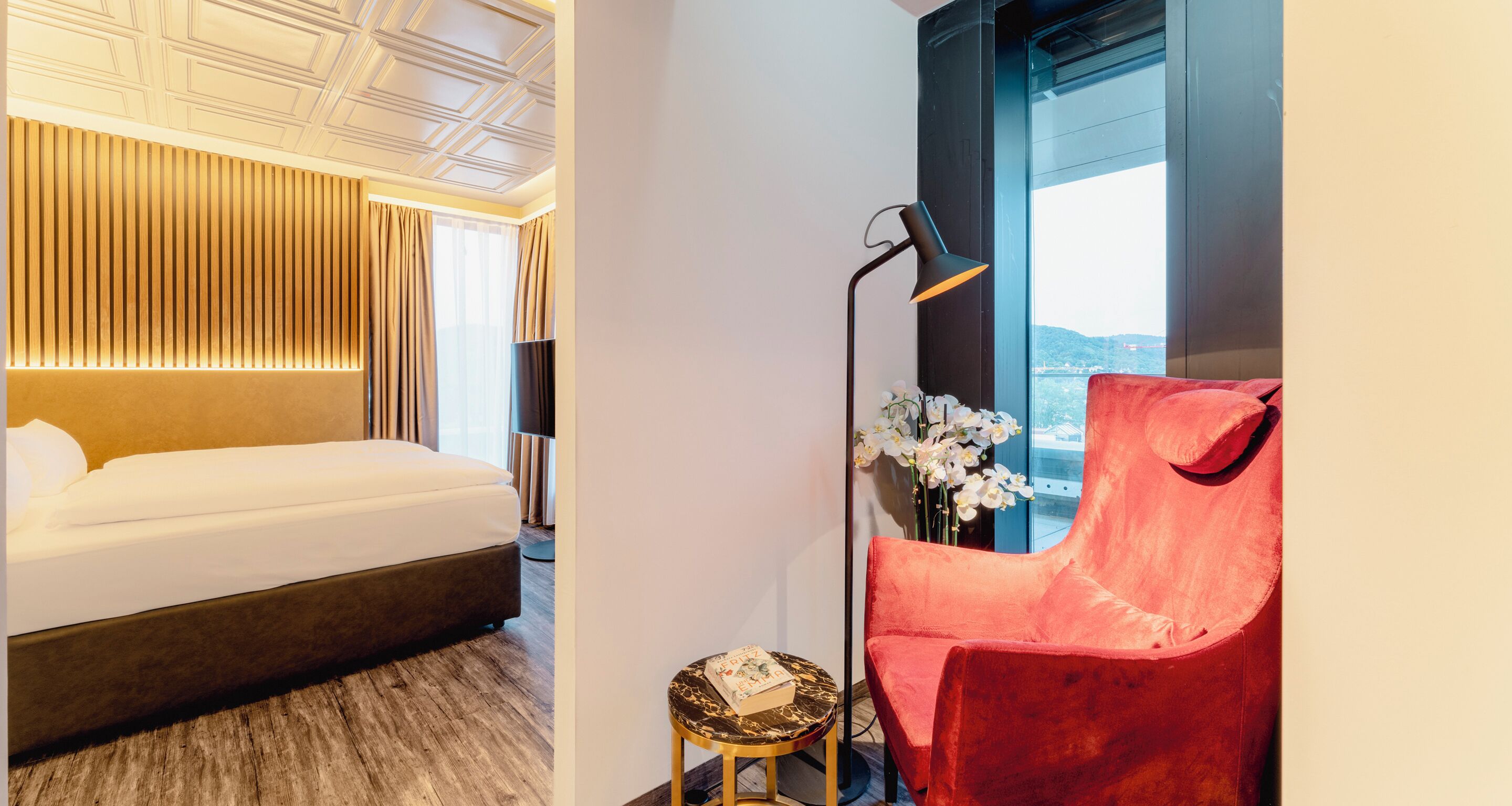 Una habitación de hotel con una cama, un sillón rojo, una lámpara de pie y una mesita con un libro.
