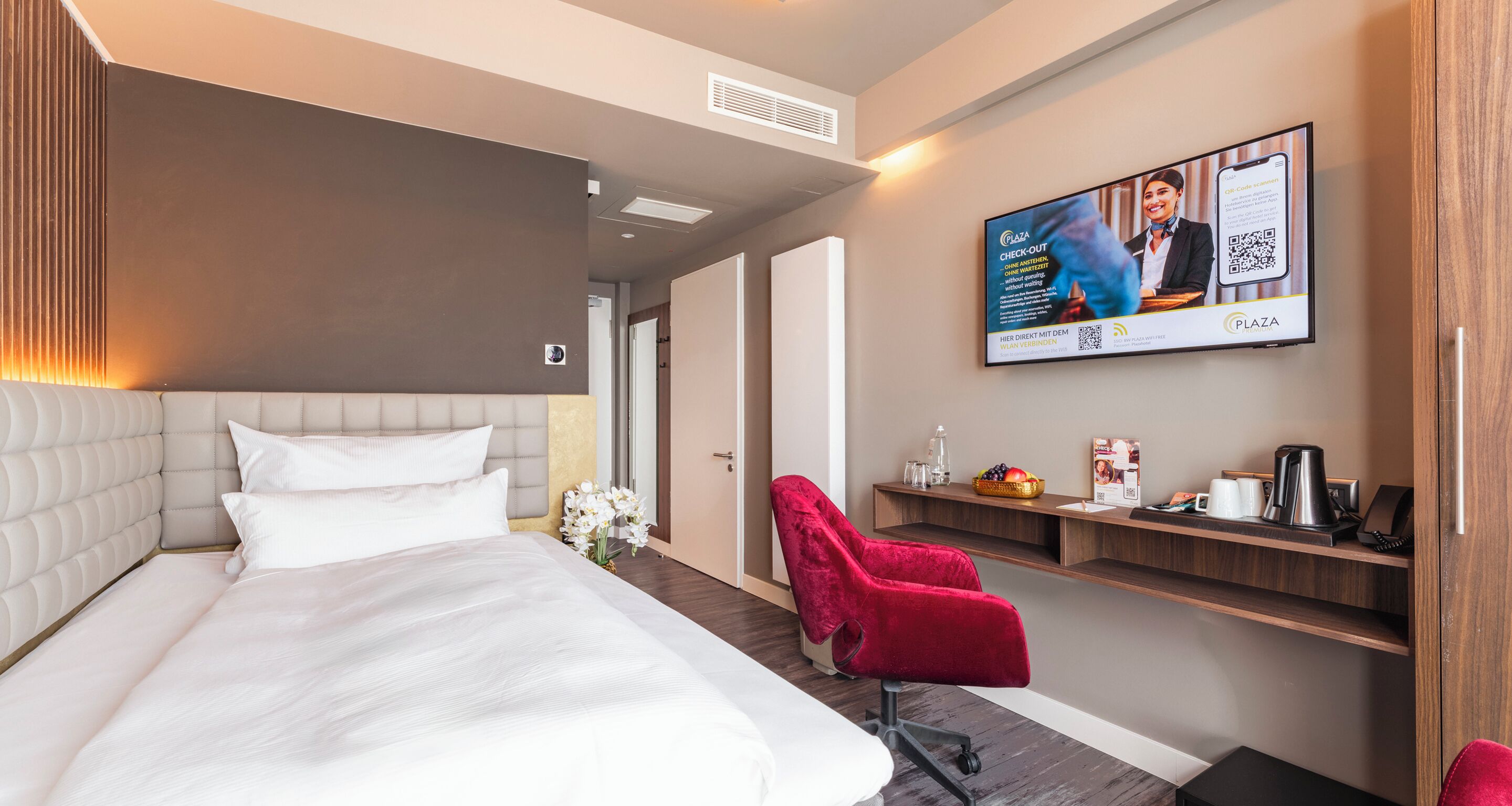 A hotel room with a large bed, a red chair, a TV screen, and a wooden shelf with amenities.