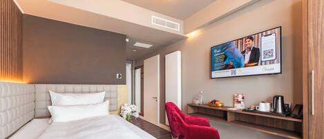 A hotel room with a large bed, a red chair, a TV screen, and a wooden shelf with amenities.