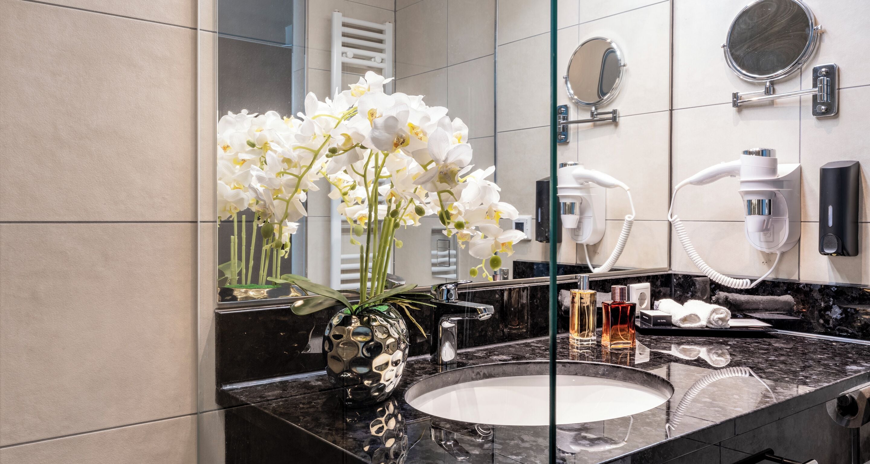 Ein Badezimmer mit Marmorwaschbecken, weißen Orchideen in einer dekorativen Vase, einem Föhn und einem Spiegel.