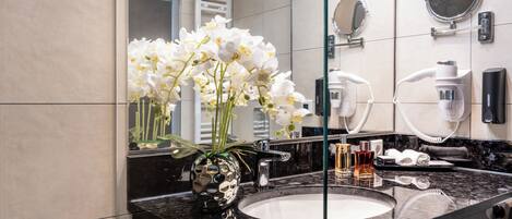 Ein Badezimmer mit Marmorwaschbecken, weißen Orchideen in einer dekorativen Vase, einem Föhn und einem Spiegel.