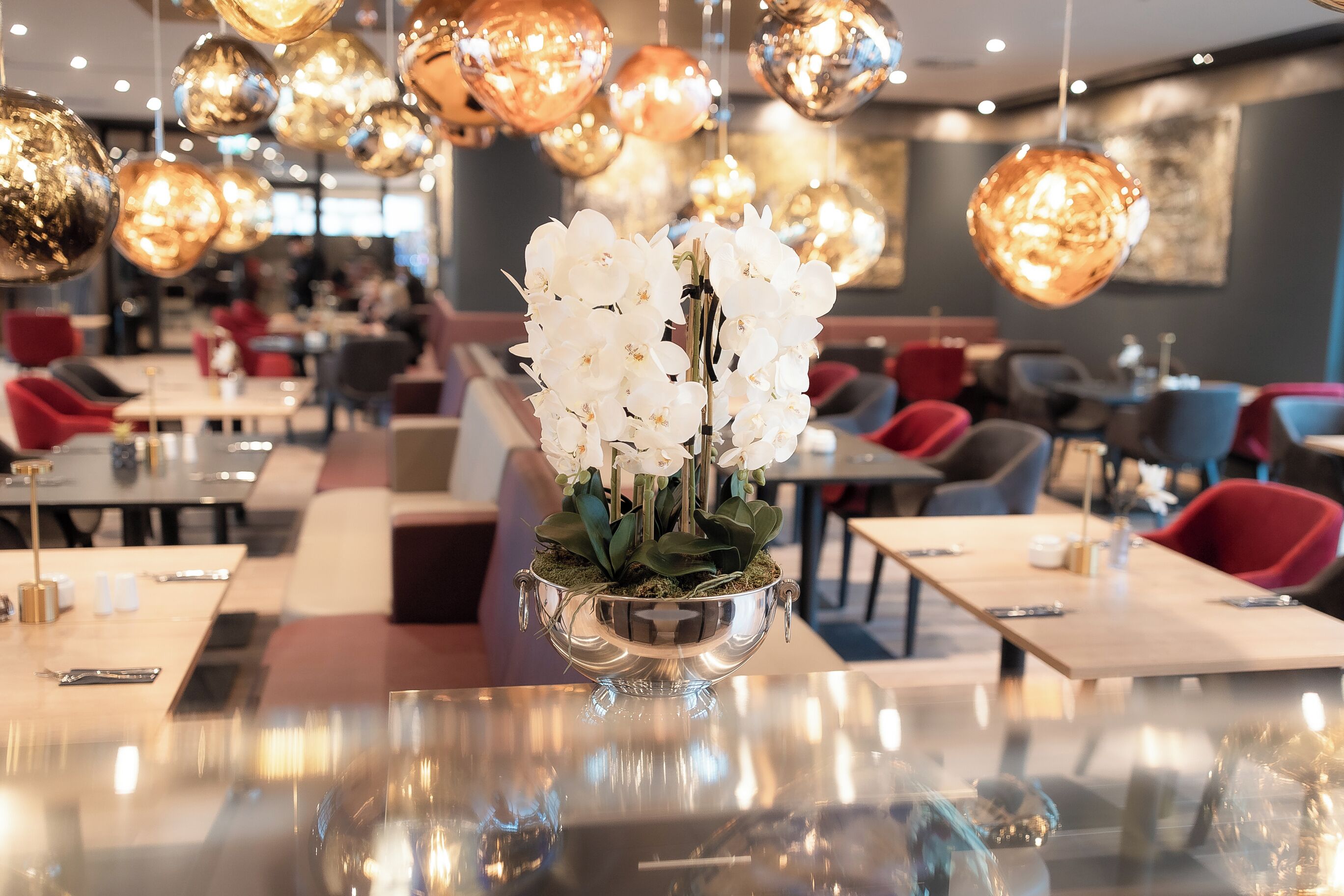 Um restaurante com mesa de vidro espelhada, um vaso de prata com flores brancas e várias luminárias pendentes esféricas.