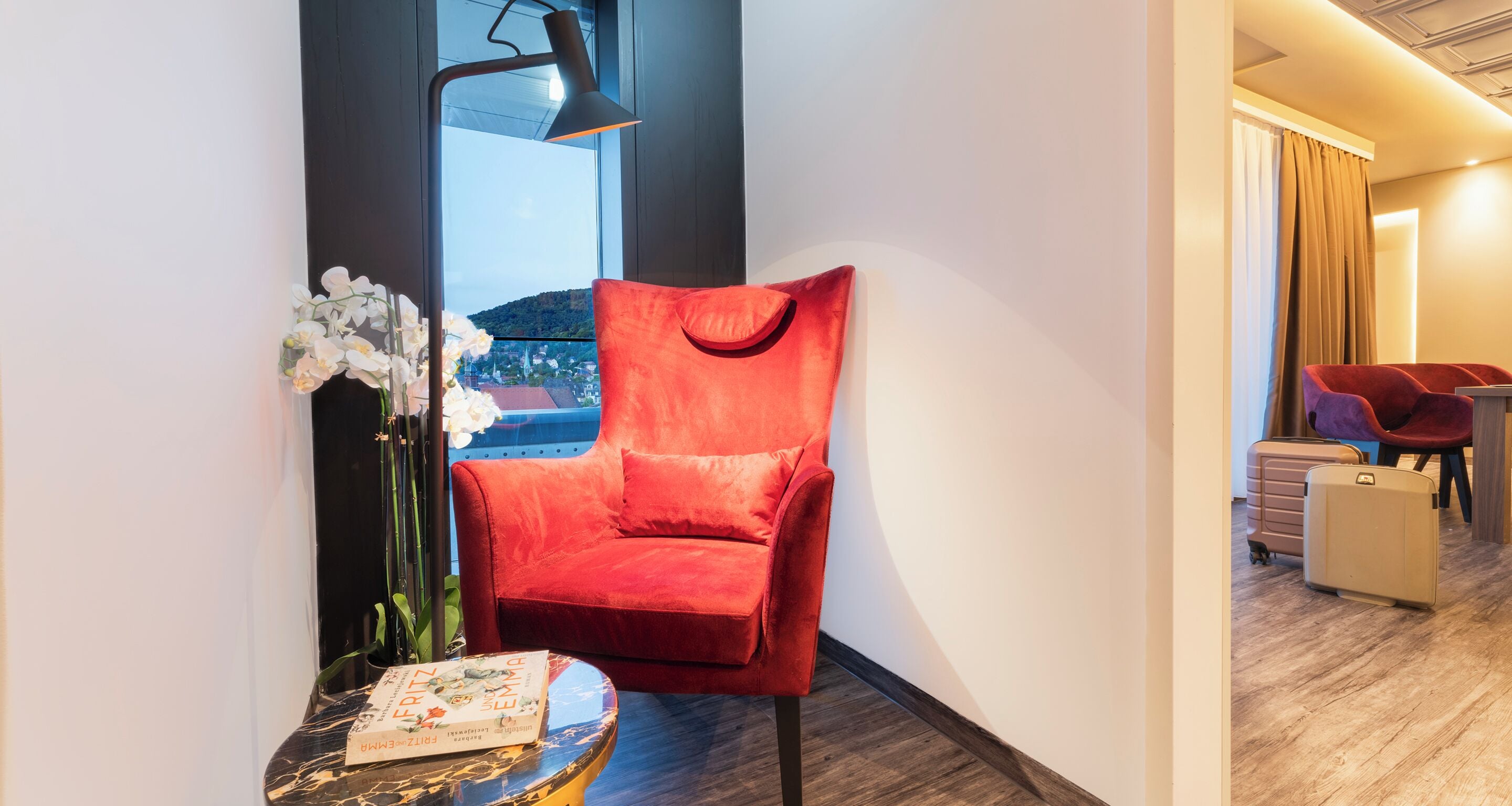 Una silla roja, una mesita con un libro y un florero con flores en una habitación de hotel.