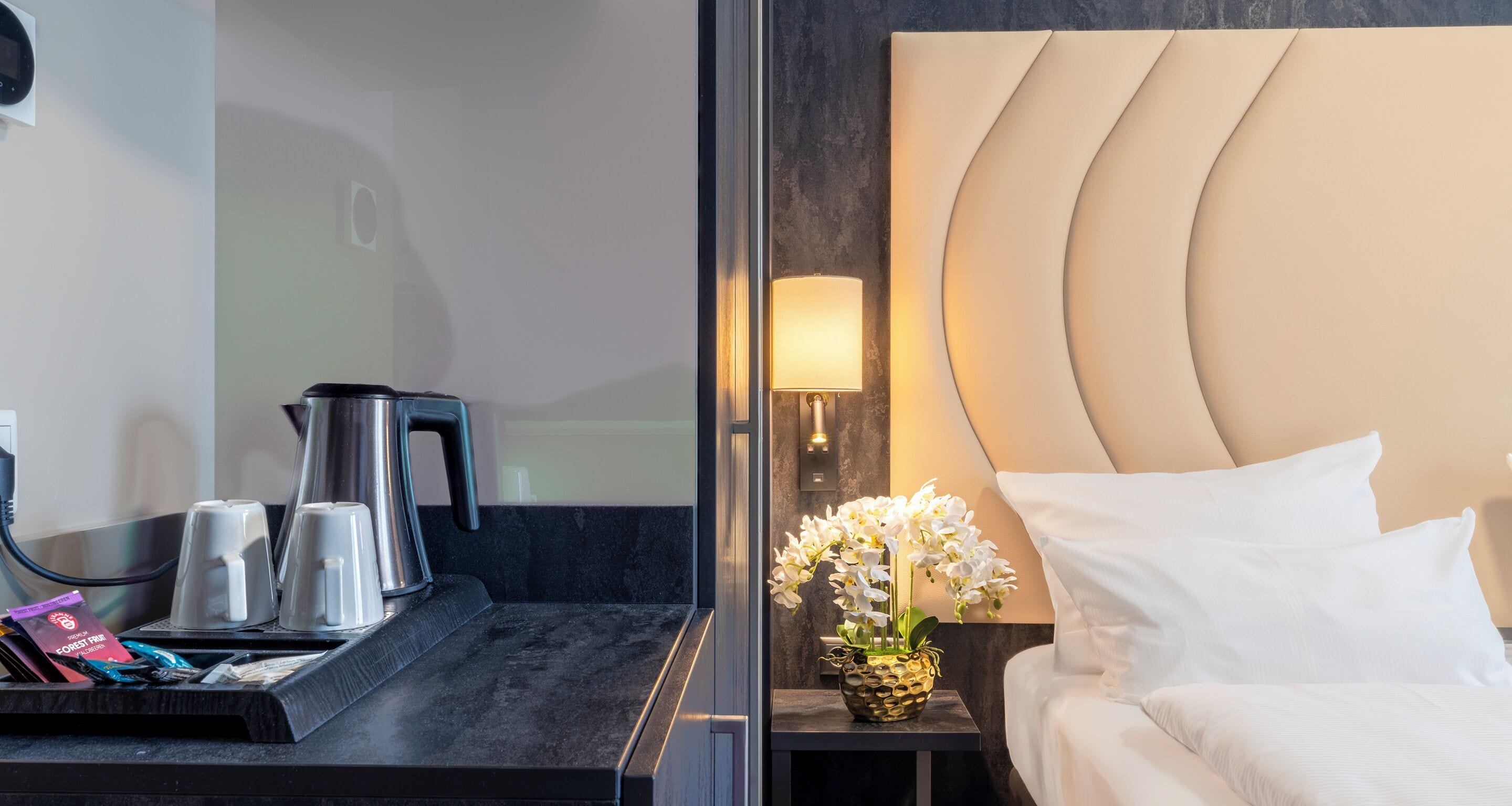 A hotel room with a bed, a bedside table with a lamp, a vase of flowers, and a coffee maker.