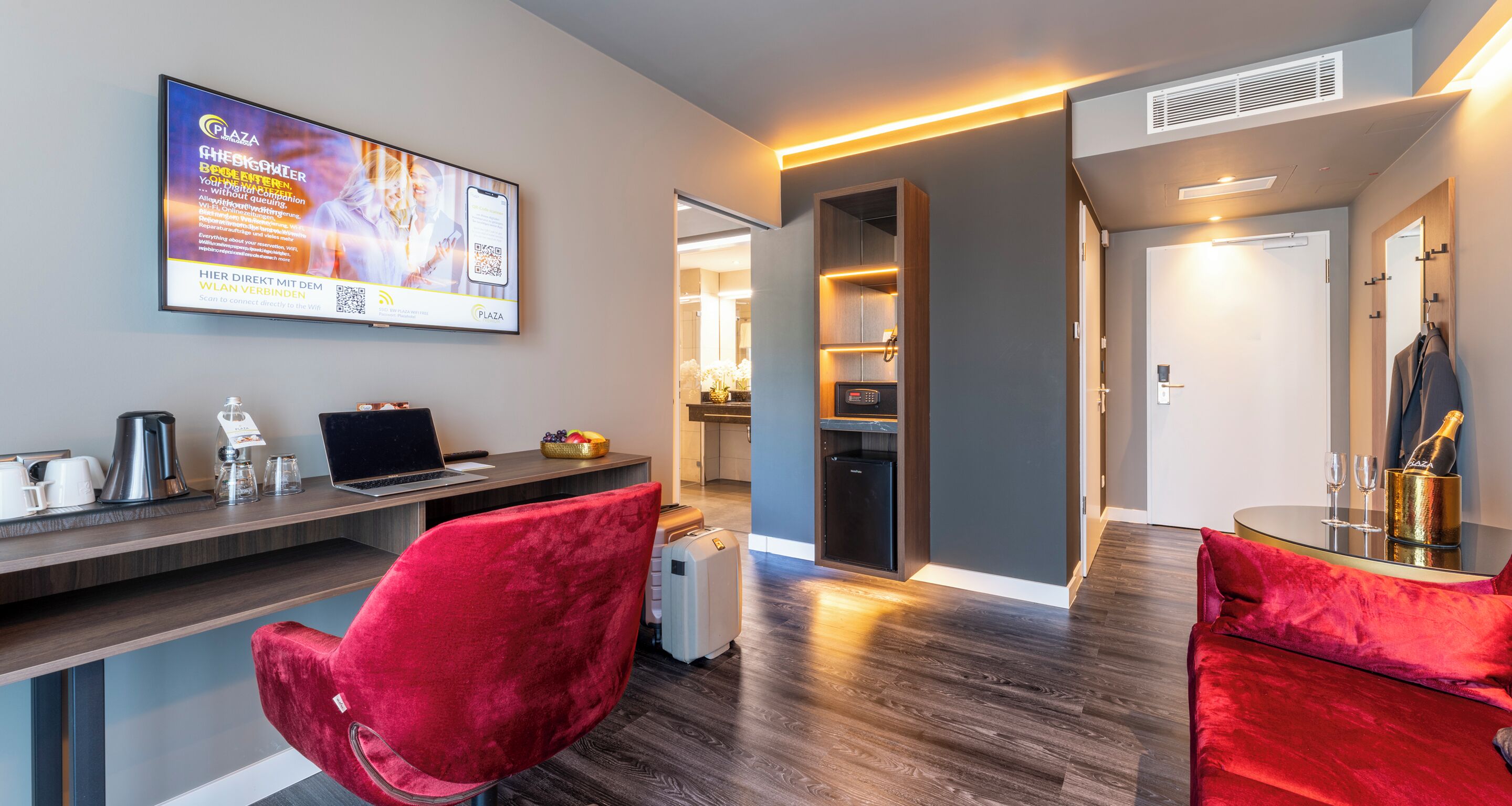 A modern hotel room with a wooden desk, a laptop, a red chair, a minibar, and a large TV screen displaying a QR code.