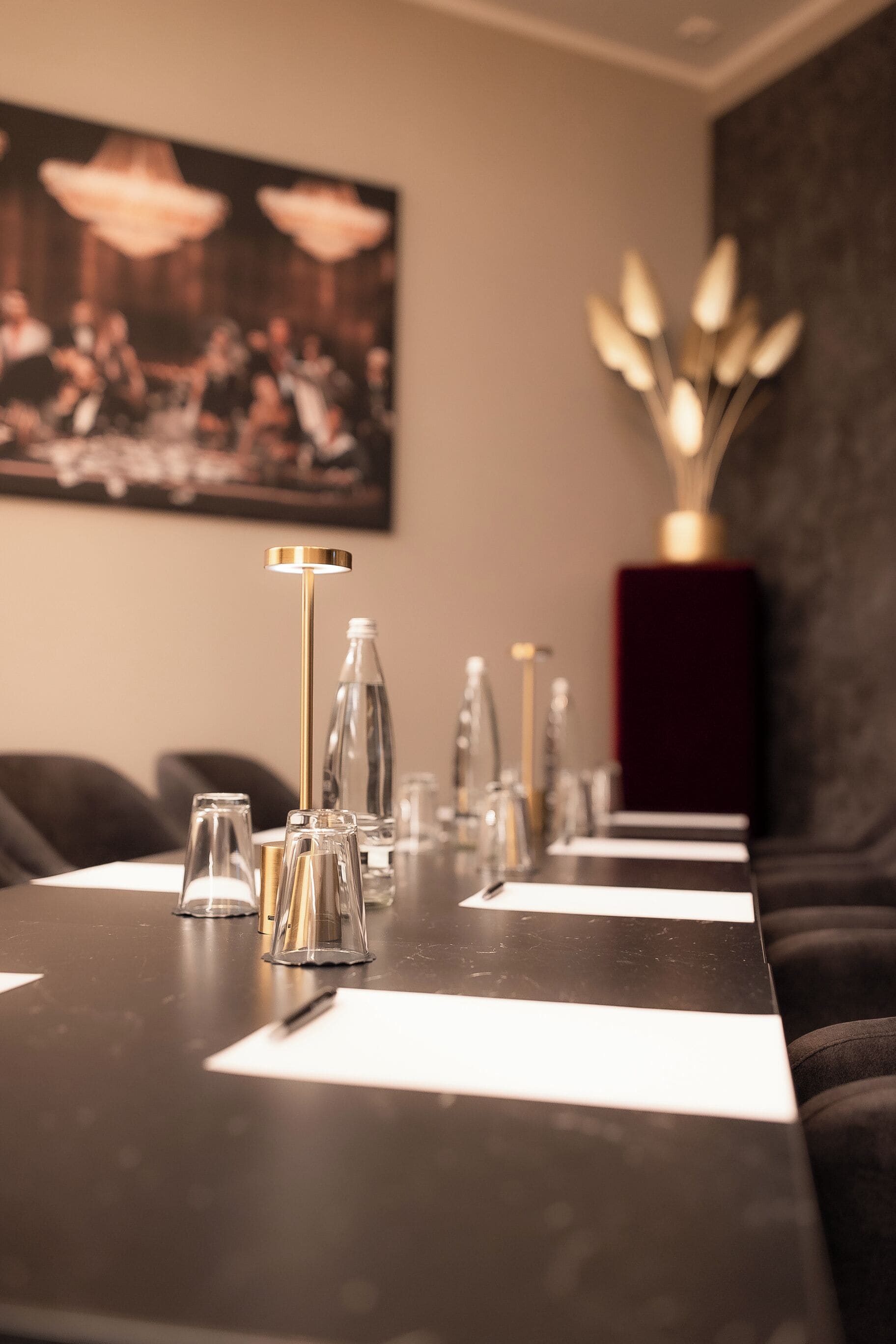Een eettafel met helder glaswerk, een goudkleurige kandelaar en een donkerrode vaas met gouden bladeren.
