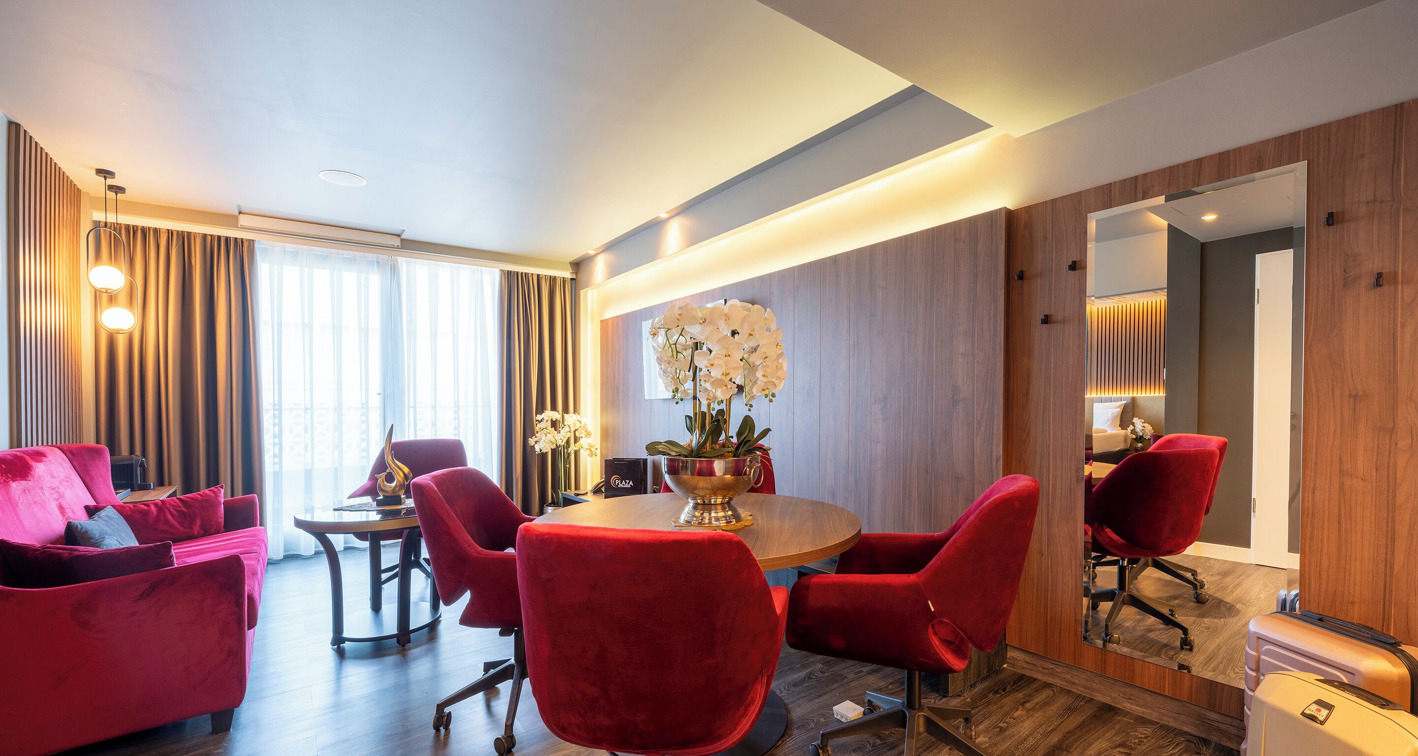 A modern hotel room with a wooden floor, red upholstered chairs, a round dining table, and a large mirror.