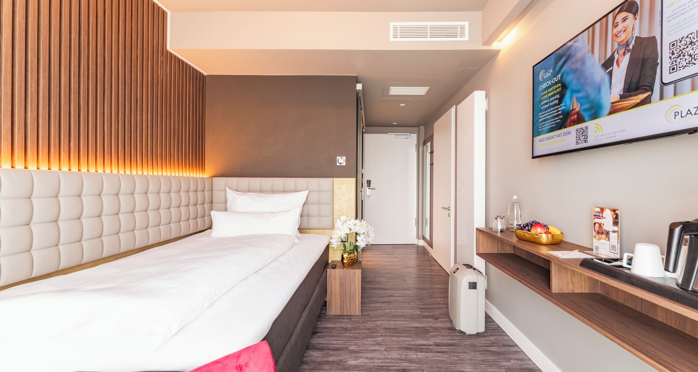 Habitación de hotel moderna con una cama grande, un cabecero de madera, una mesita de noche de madera con un florero y una maleta blanca, una cafetera y un televisor empotrado mostrando un anuncio.