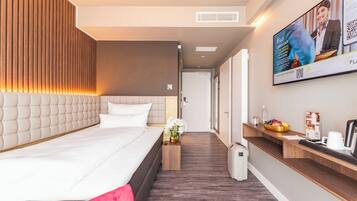 Habitación de hotel moderna con una cama grande, un cabecero de madera, una mesita de noche de madera con un florero y una maleta blanca, una cafetera y un televisor empotrado mostrando un anuncio.