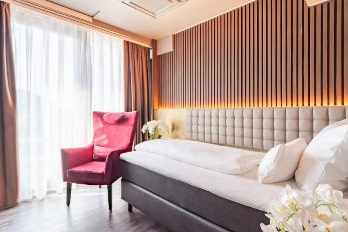 A hotel room with a bed, a red chair, a window with curtains, and a wall with vertical wooden slats.