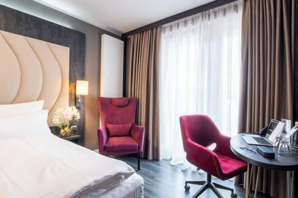 A hotel room with a bed, a desk with a laptop, a red chair, and a lamp.
