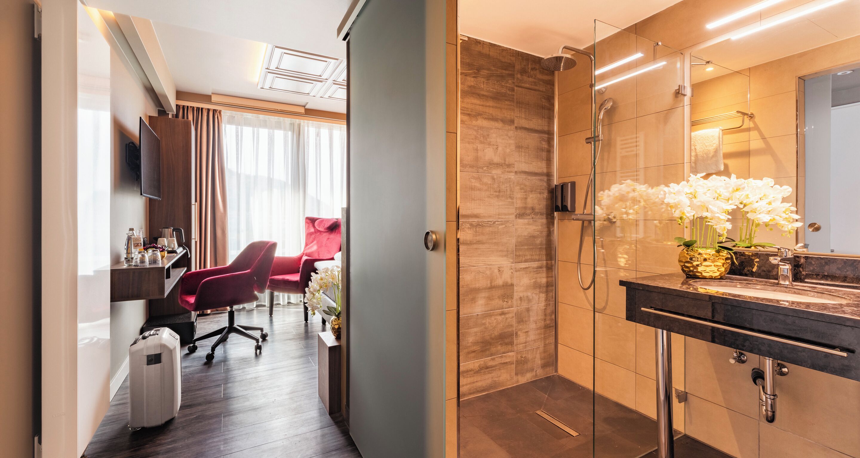 Ein modernes Hotelbad mit einer in Glas eingefassten Dusche, einem Waschtisch mit Spiegel sowie einem Schreibtisch mit Stuhl und einem roten Sessel.