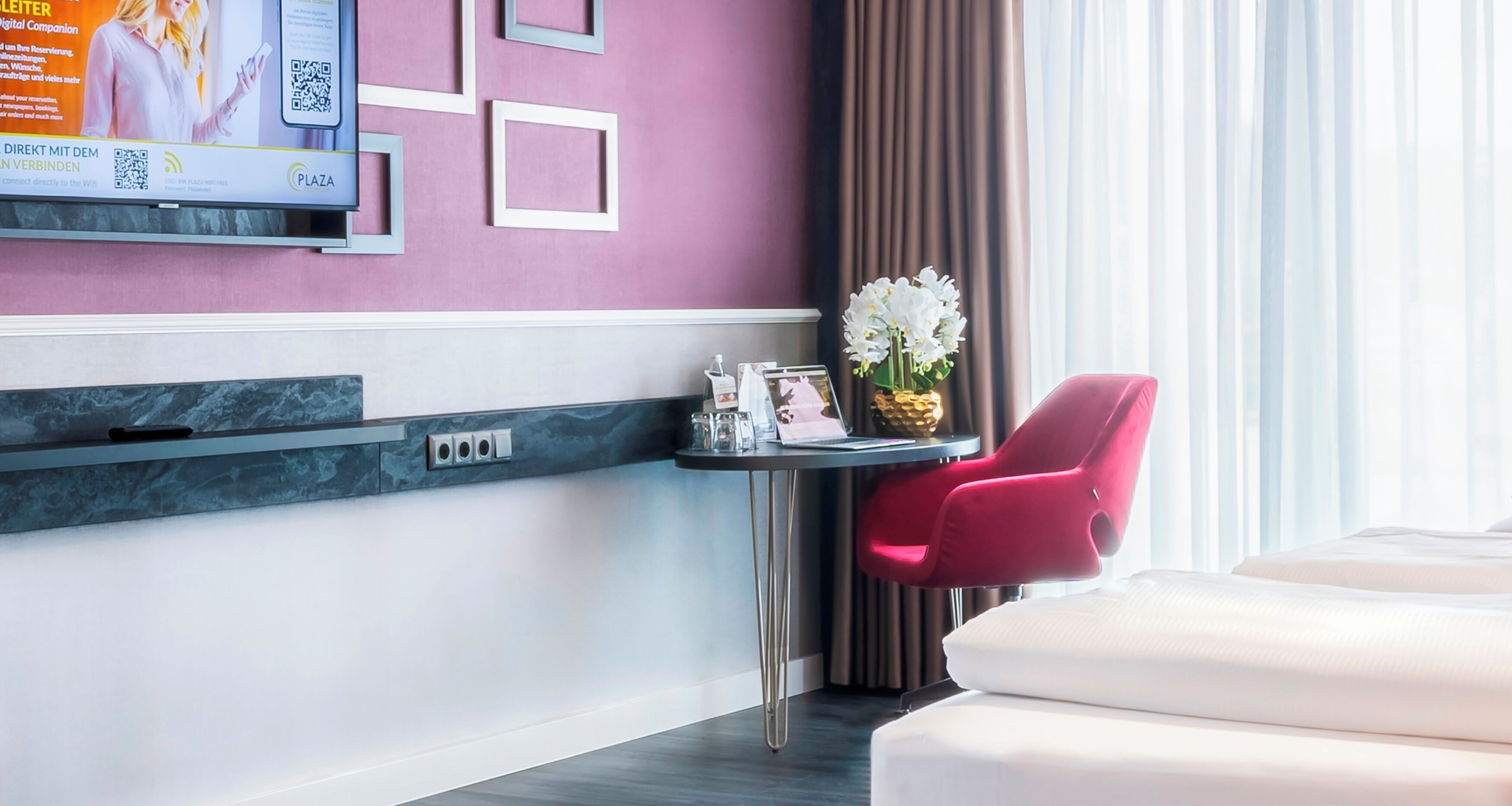 Habitació d'hotel amb un llit, una cadira vermella, una taula petita amb un vas de flors, un televisor penjat a la paret i una paret decorativa morada.