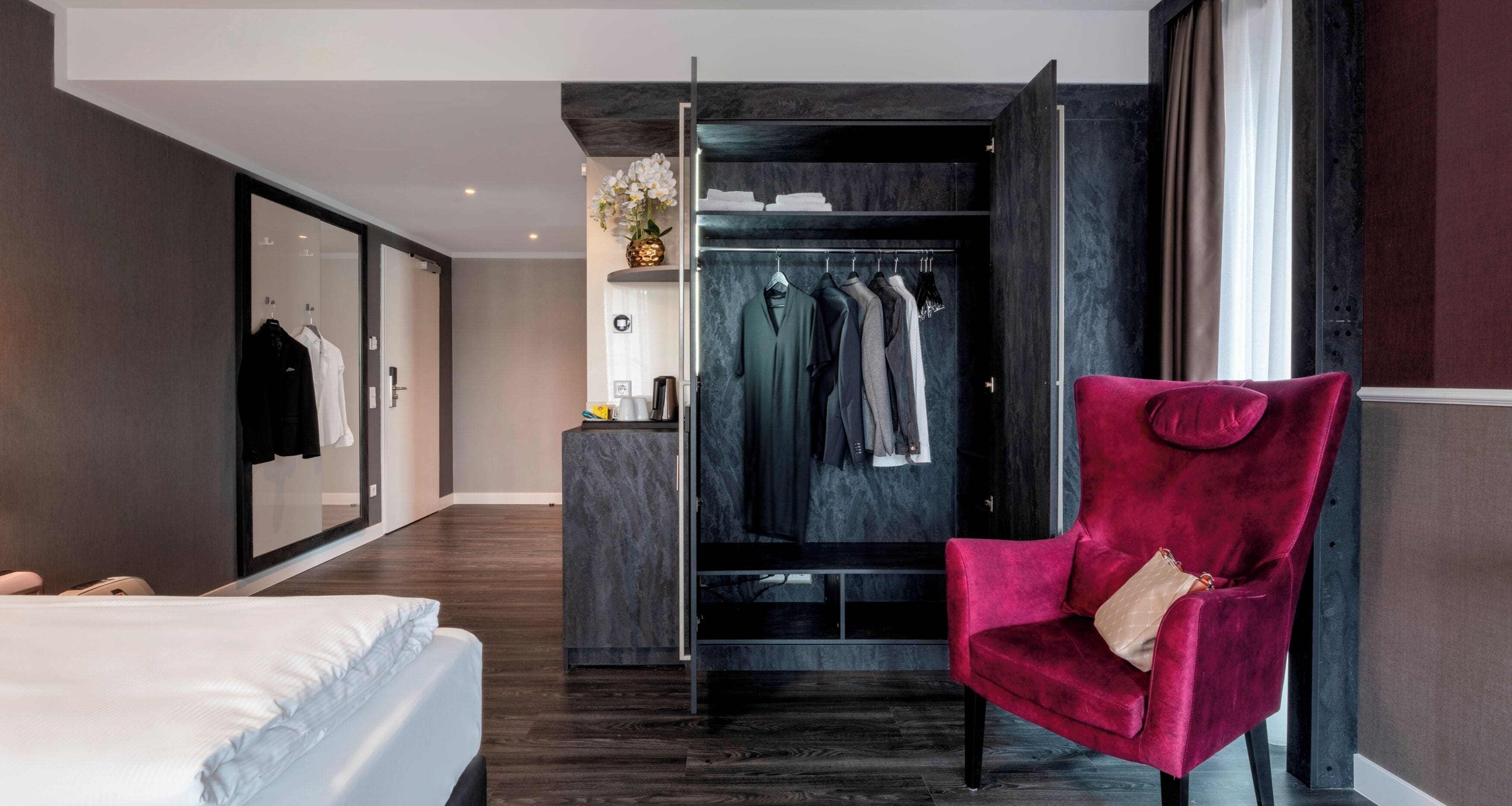 Ein modernes Hotelzimmer mit einem großen Kleiderschrank, einem roten Samtstuhl und einem Bett.
