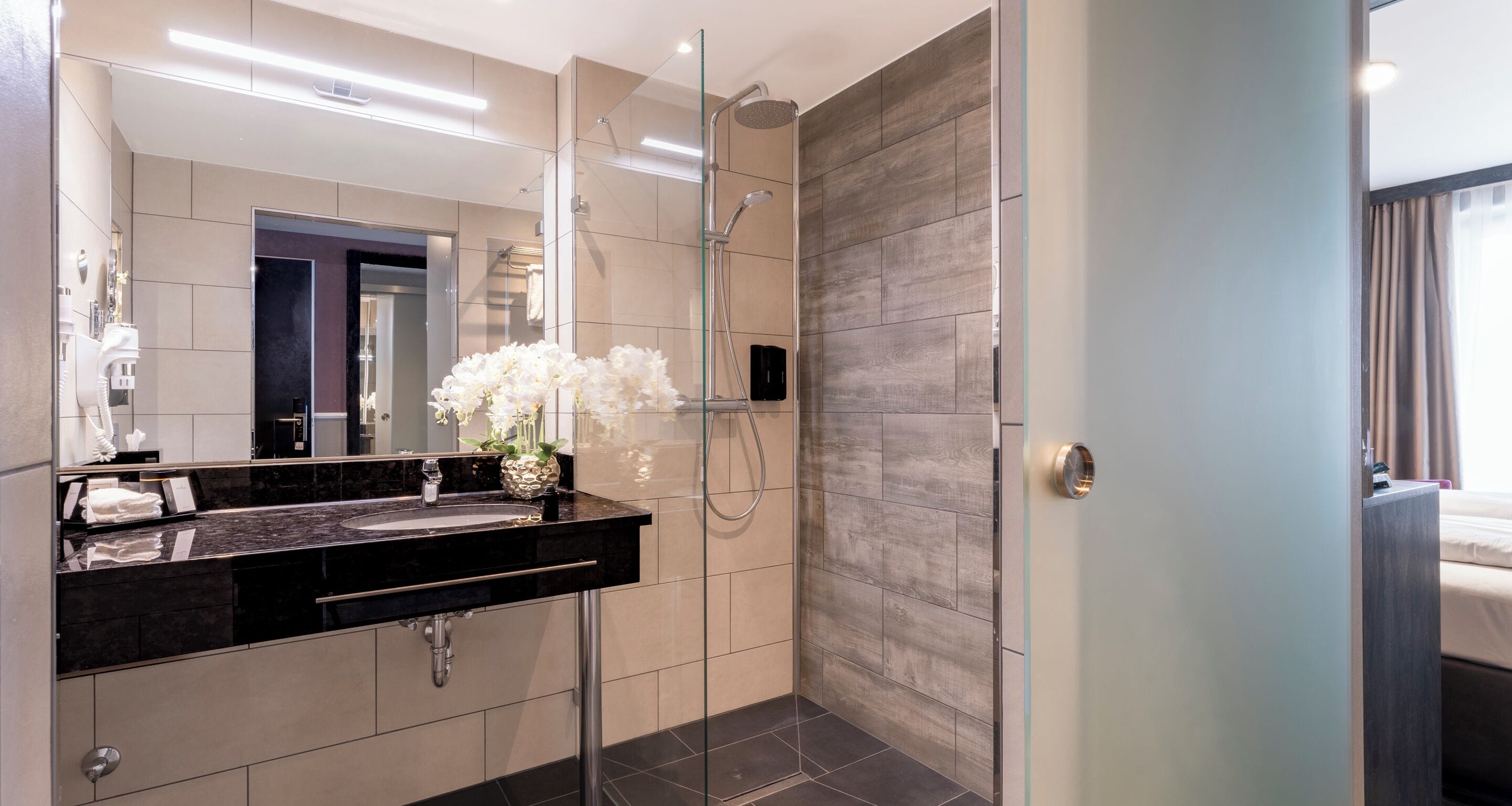 A modern bathroom with a black countertop sink, a glass shower enclosure, and a mirror with a decorative vase of white flowers.