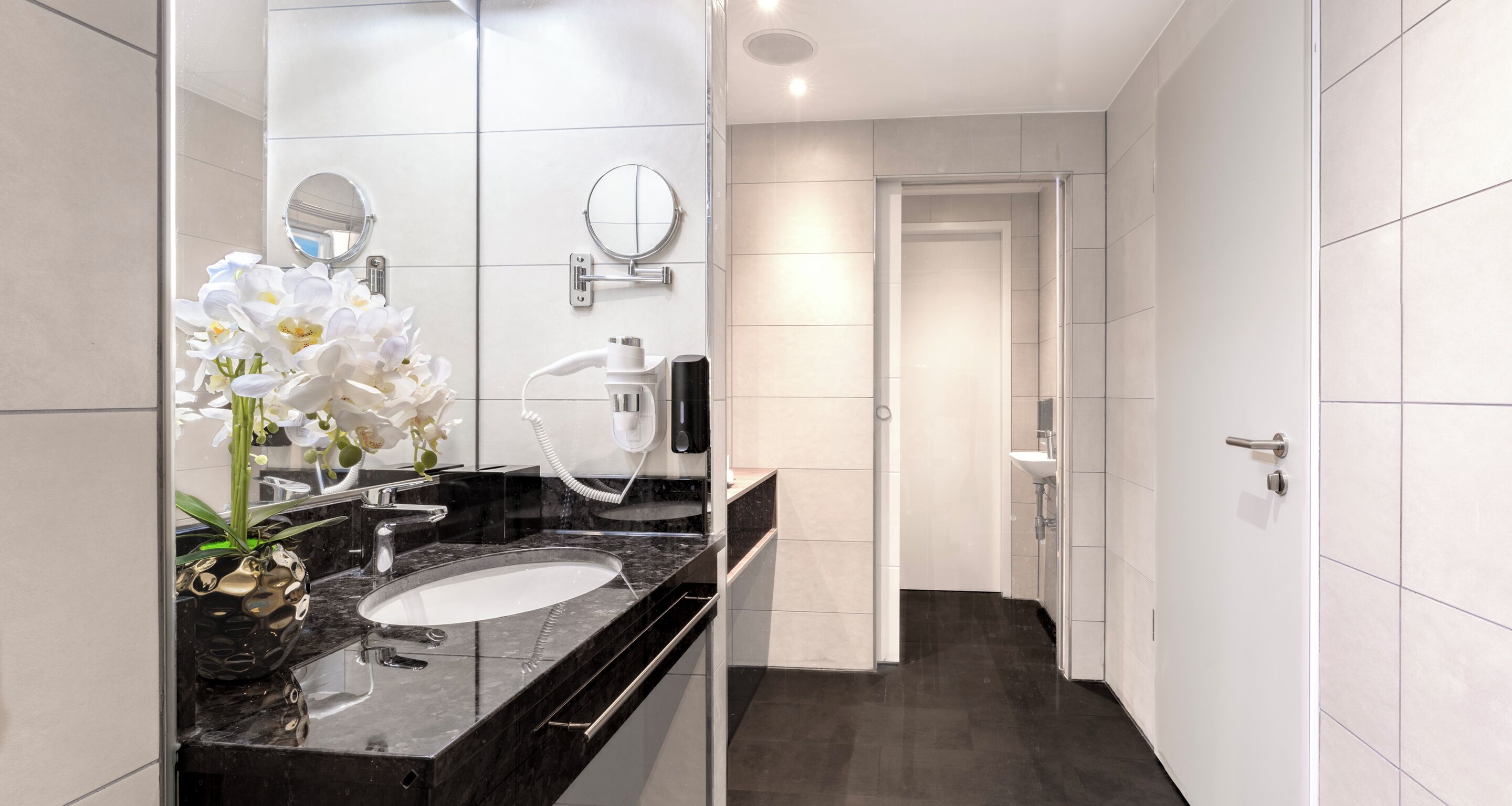 A modern bathroom with a black marble sink, white tiles, a round mirror, and a white orchid plant.