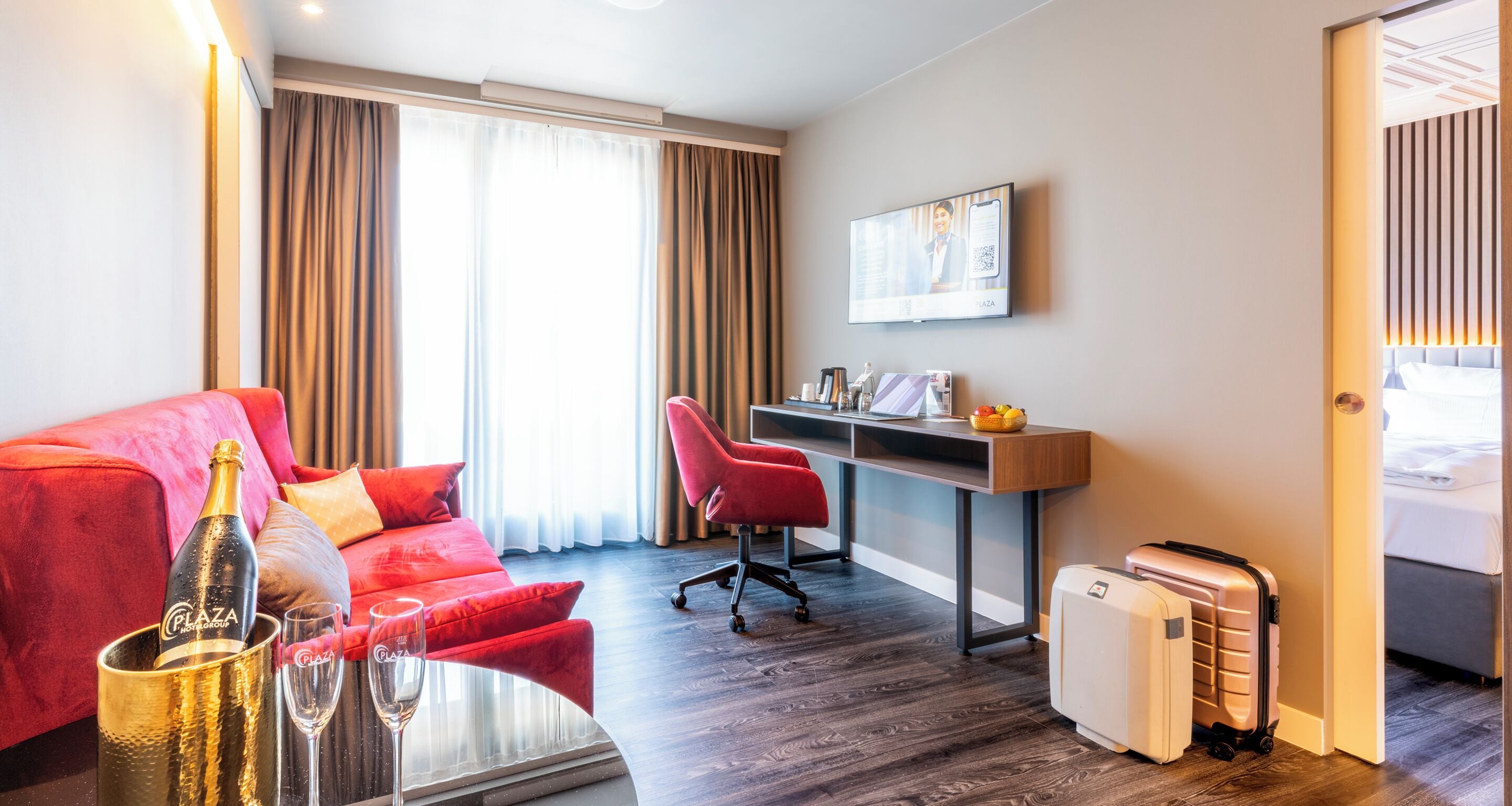 A hotel room with a red sofa, a desk, a TV, a minibar, and a suitcase.