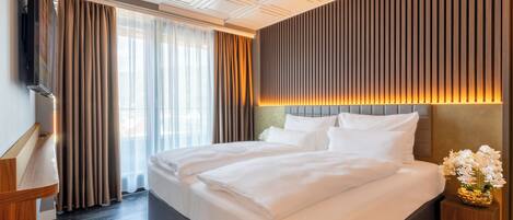A hotel room with a large bed, a flat-screen TV, a wooden nightstand, and a vase of flowers.