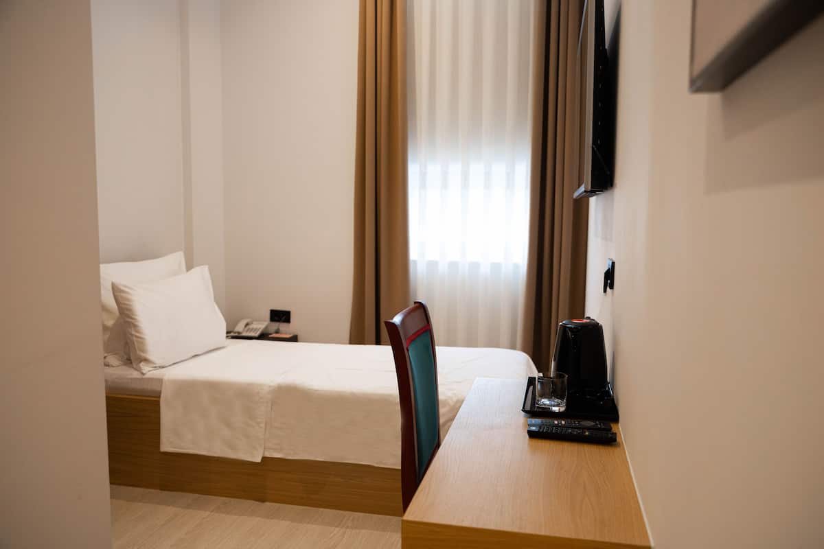 A hotel room with a bed, a wooden desk, a chair, a TV mounted on the wall, and a window with curtains.