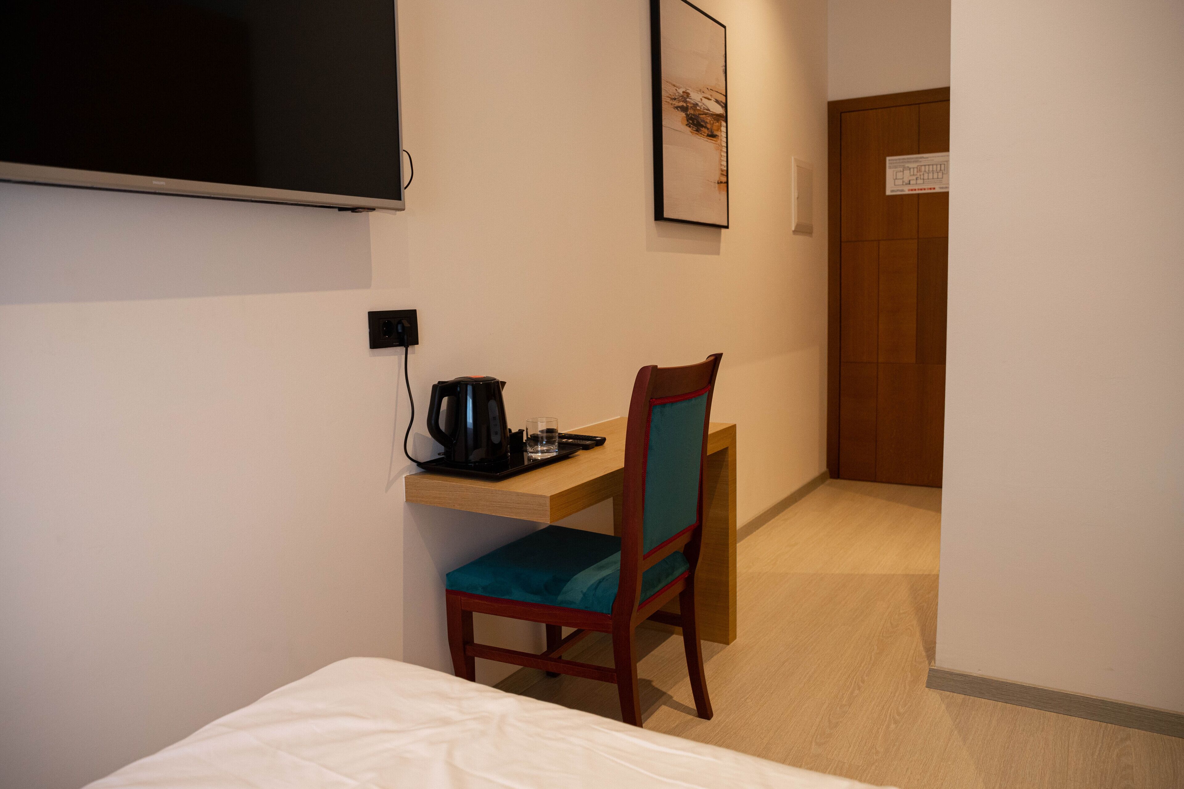Habitación de hotel con una cama, un escritorio con una silla, un televisor montado en la pared y una puerta al fondo.