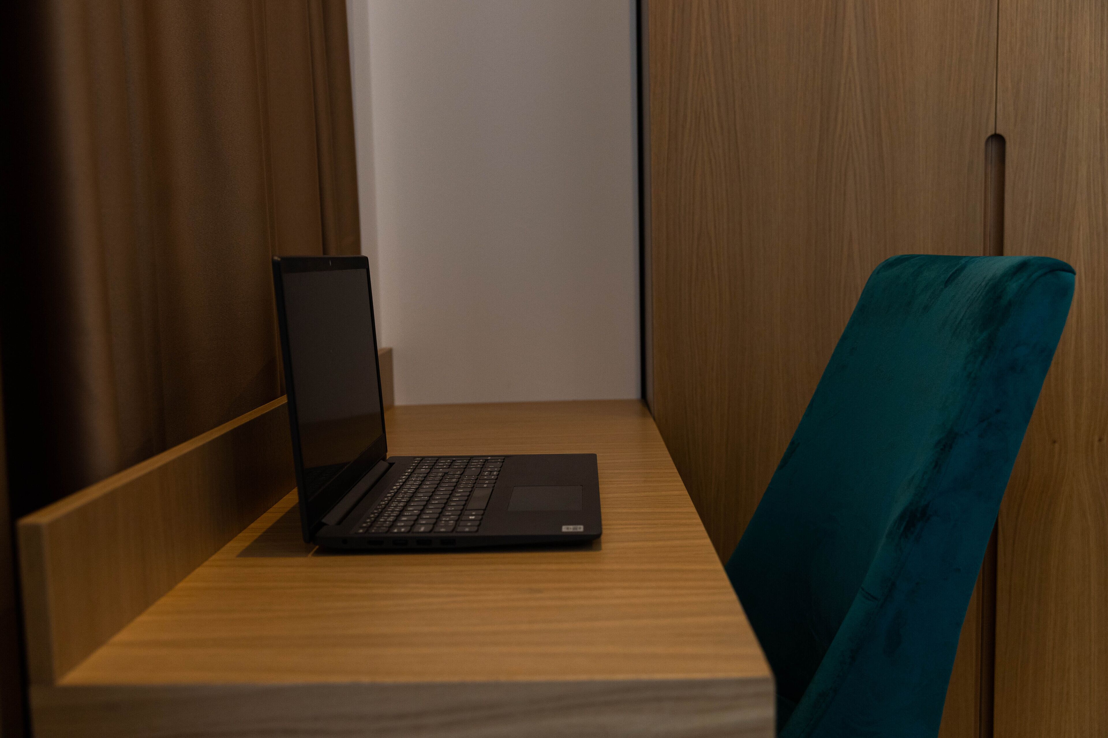 A laptop on a wooden desk in a room with a teal chair.