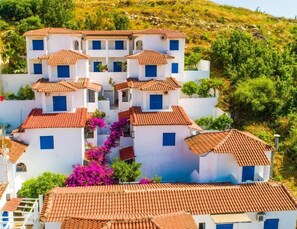 A multi-story building with terracotta tiled roofs and white walls, surrounded by greenery and hills.