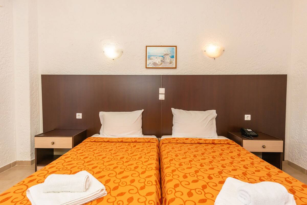 Kahden hengen huone, jossa on kaksi sänkyä. Jokaisessa sängyssä on oranssikuviollinen peitto ja valkoiset tyynyt. Huoneessa on kaksi yöpöytää, joissa on lamput, ja sängyn päädyn yläpuolella on pieni taulu.