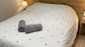 A bedroom with a wooden headboard, a white bedspread with a tree pattern, a rolled towel, a bedside lamp, and an air conditioning unit. - CHALET ROMANE, SLEEPS 6, LAKE VIEW, AIR-CONDITIONED, BALCONY, MOTORCYCLE ROOM, GERARDMER (Gérardmer)