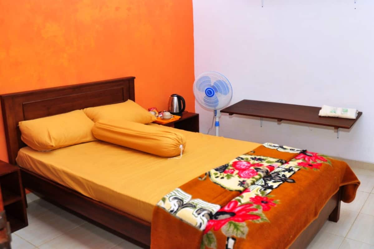 Una cama con una colcha amarilla y un edredón floral, una mesita de noche con una tetera, un ventilador y un estante con un teléfono.