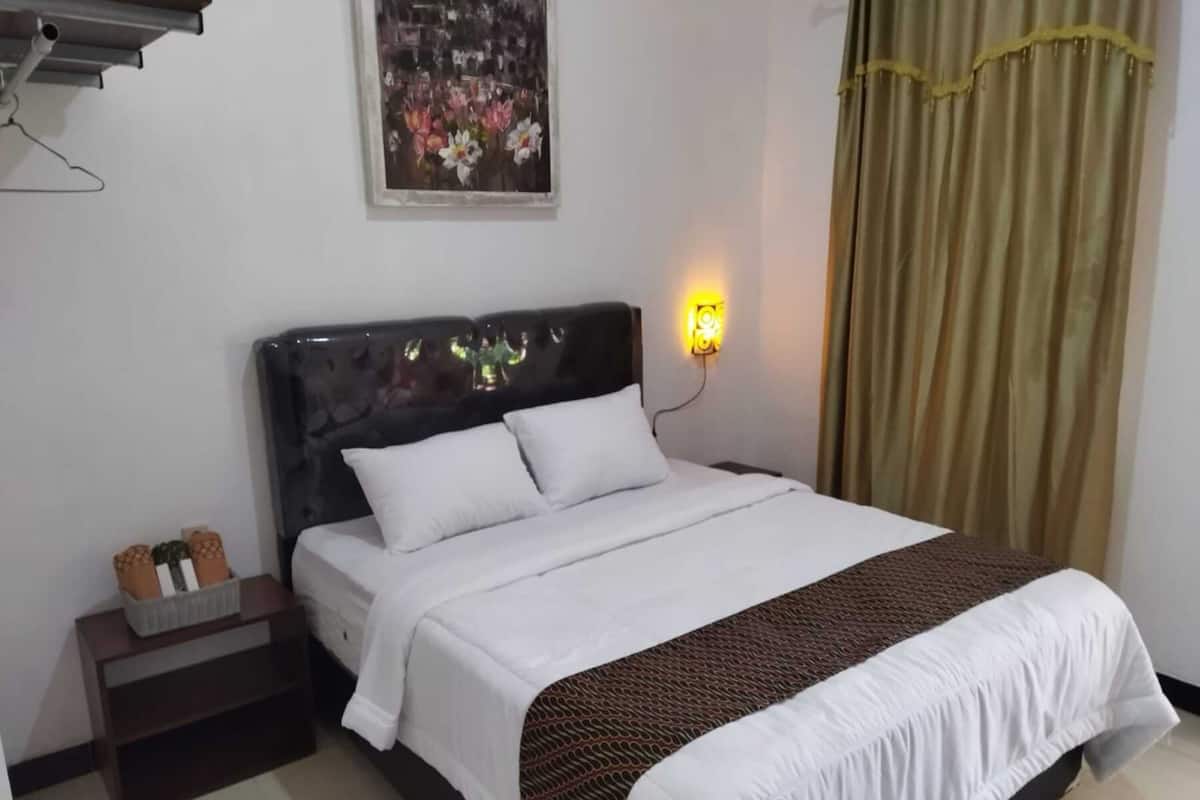 Una cama bien tendida con ropa blanca y un plaid estampado, una mesita de noche con un cesto de artículos de tocador, una lámpara de pared y un cuadro de flores sobre la cama.