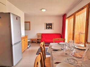 Cozinha compacta com geladeira, armários de madeira, uma mesa de jantar posta para dois e um sofá vermelho ao fundo.