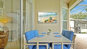 A dining area with a table set for four, blue cushioned chairs, a small potted plant, and a beach-themed painting on the wall. - Windsor Hills! Free Pool Heat/BBQ/Waterpark. Game Room w/Theater, Themed Rooms. (Kissimmee)