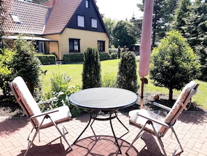 Een terras met een ronde tafel en twee stoelen, omgeven door een tegelpad en begroeiing.