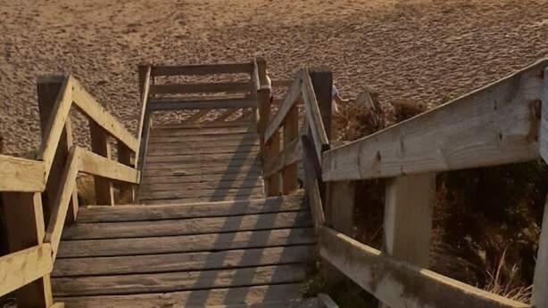 Eine Holztreppe führt zu einem Strand, im Hintergrund versinkt die Sonne im Abendrot.