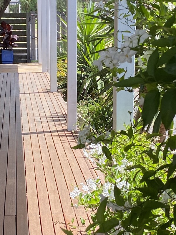 Une terrasse en bois, agrémentée de piliers blancs, entourée de verdure et de fleurs blanches.