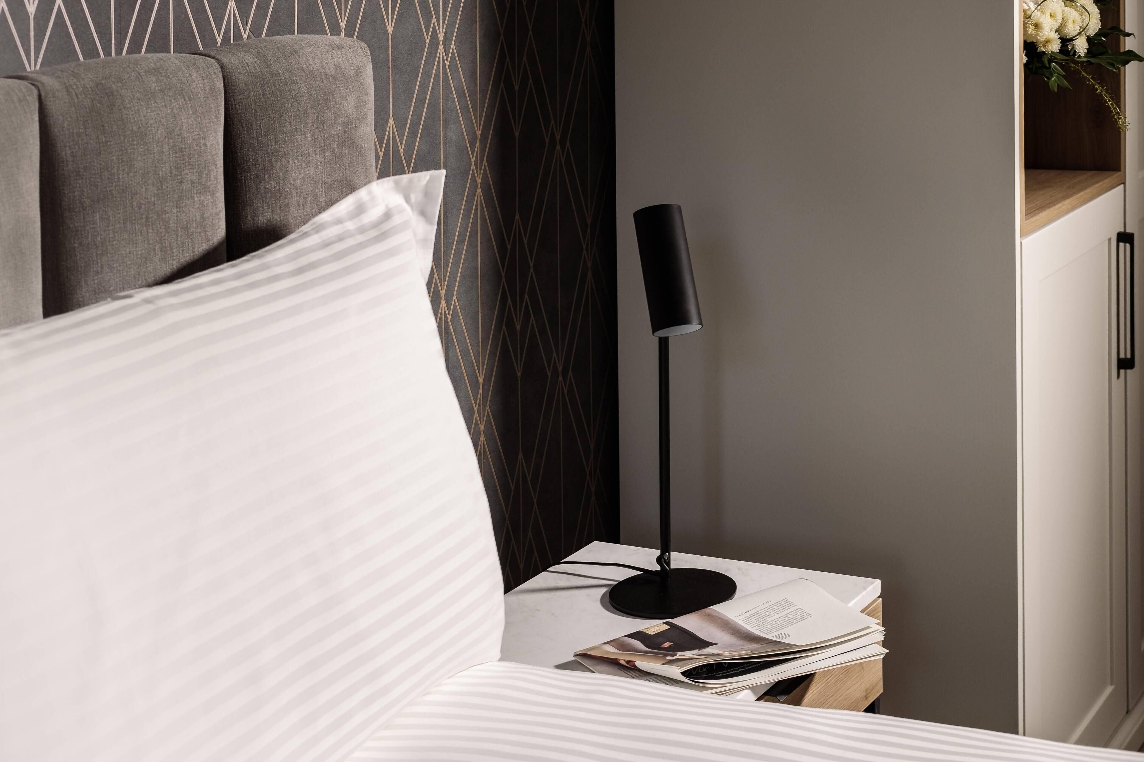A neatly arranged bedroom with a bed, a bedside table with a lamp, and a wall with a geometric pattern.
