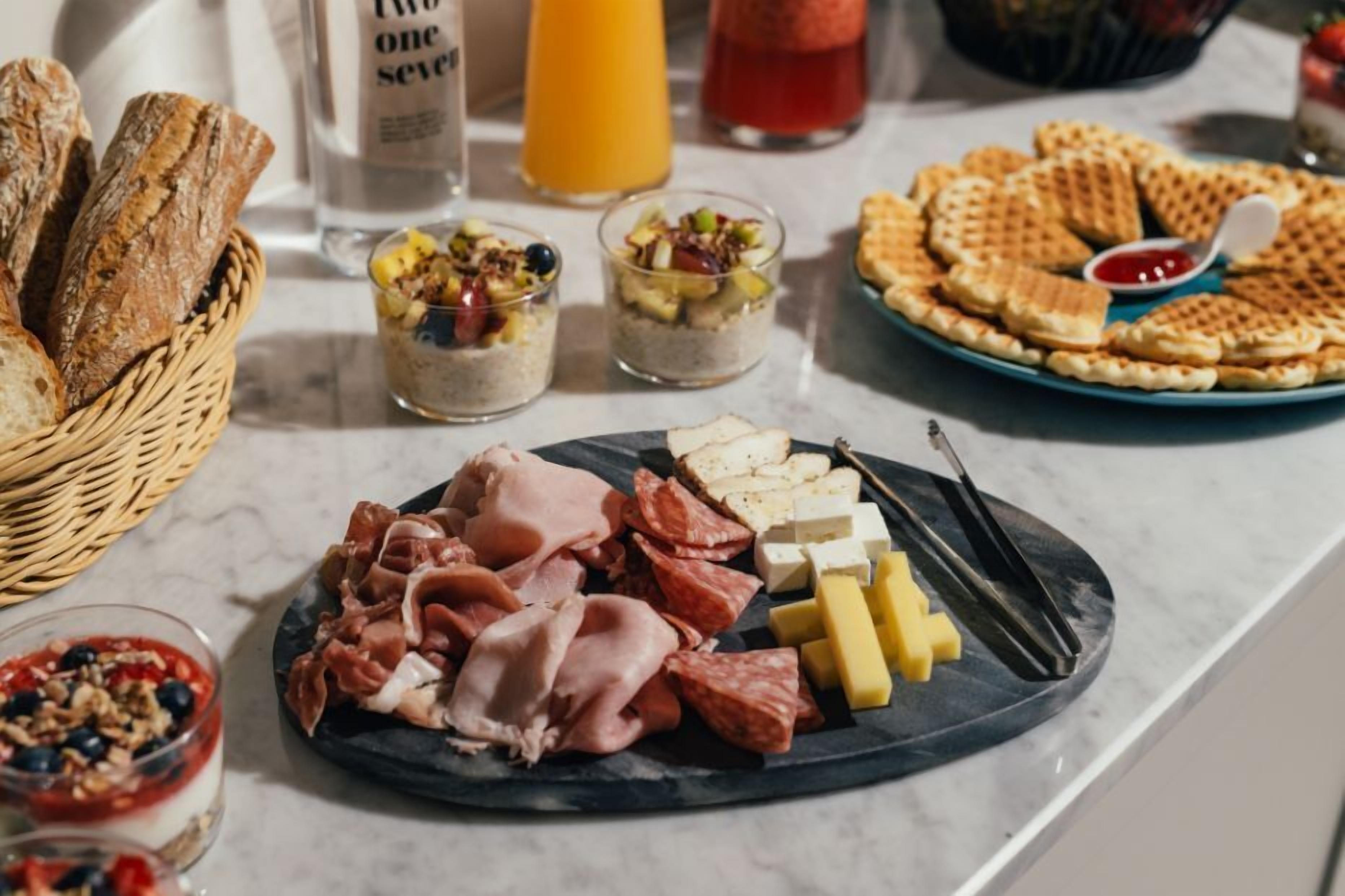 A breakfast spread with bread, cheese, meats, and waffles on a marble countertop.